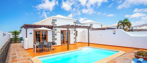 A swimming pool with a tiled deck, a covered patio area, and a white building with a tiled roof.