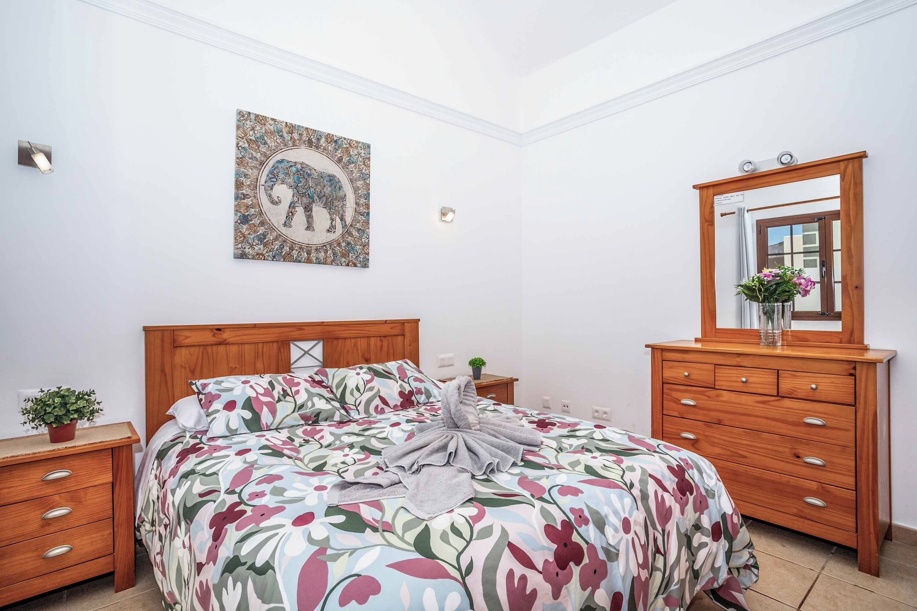Un dormitorio con cubrecama floral, cabecera de madera, mesita de noche con planta, espejo e imagen colgada en la pared de un elefante.