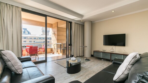 A modern living room with a glass coffee table, leather sofas, a flat-screen TV, and a sliding glass door leading to an outdoor dining area. - Raphael Penthouse | Square and Mall and Pool Access (Johannesburg)