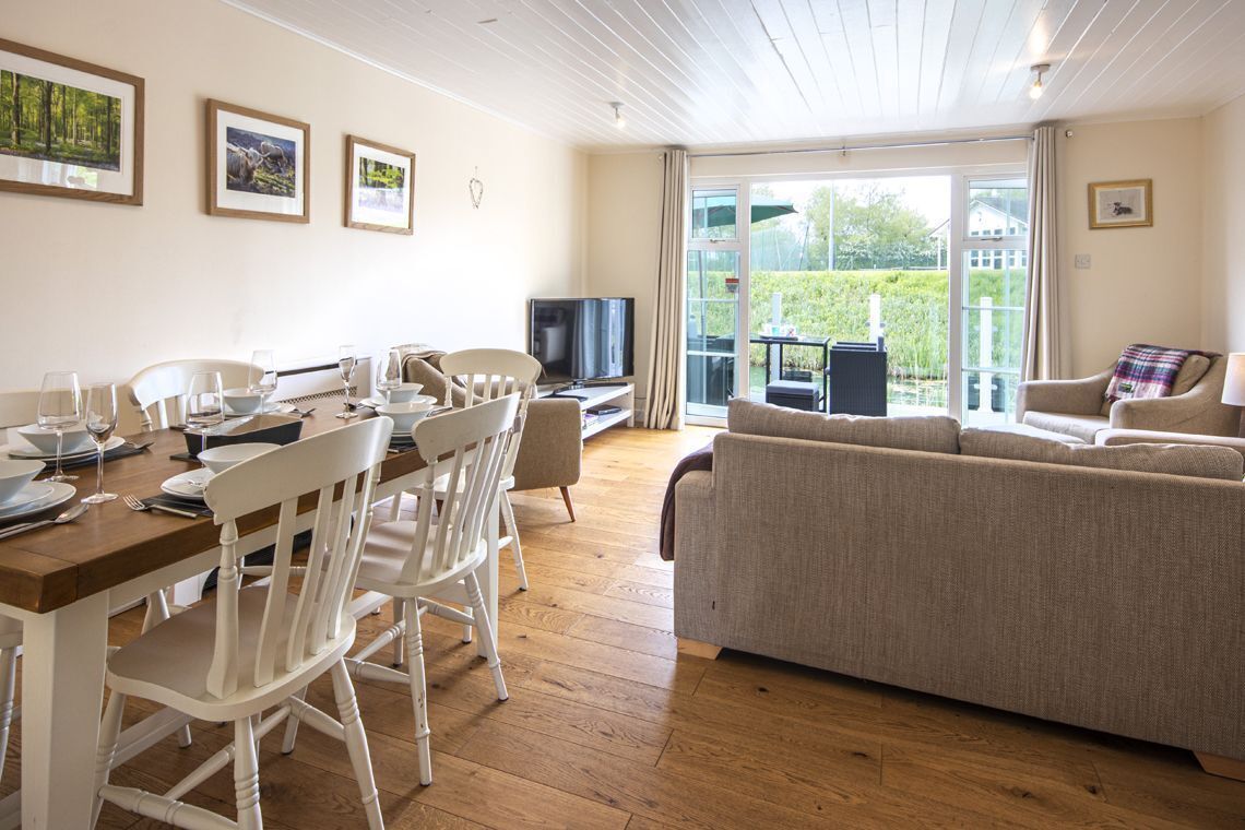 A dining area with a wooden table set for four, white chairs, and a television on a stand. There is a sofa and a chair in the living area with a view of greenery outside.