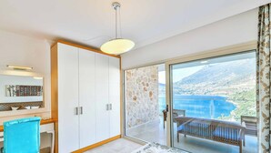Ein Hotelzimmer mit Schreibtisch, Stuhl, Kleiderschrank, einem Balkon mit Sitzbank und Blick auf eine gebirgige Küstenlandschaft.