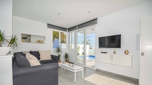 A modern living room with a grey sofa, a coffee table, a TV mounted on the wall, and a sliding glass door leading to a pool.