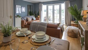 A cozy living room with a sofa, a dining table set for two, and a large window with curtains. - Windrush Lake 87, The Retreat (P) - Lakeside Lodge, On-site Facilities, Sleeps 6 (South Cerney)