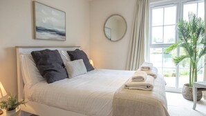 A neatly made bed with a white headboard, a bedside table with a lamp, a large window with curtains, and a potted plant. - Windrush Lake 87, The Retreat (P) - Lakeside Lodge, On-site Facilities, Sleeps 6 (South Cerney)