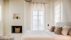 A minimalist bedroom with a large bed, a small bedside table, a wall-mounted shelf, and a window with sheer curtains.