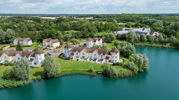 A lakeside residential area with multiple houses, some with towers, surrounded by greenery and a calm lake. - Windrush Lake 88, Waterside - Lakeside Lodge, On-site Activities, Sleeps 8 (SOUTH CERNEY)