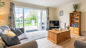 A living room with a sofa, coffee table, TV, bookshelf, and sliding glass doors to a garden. - Windrush Lake 88, Waterside - Lakeside Lodge, On-site Activities, Sleeps 8 (SOUTH CERNEY)