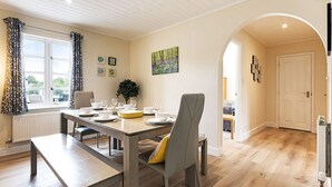 A dining area with a table set for four, a potted plant, and a view of the outdoors through a window. - Windrush Lake 88, Waterside - Lakeside Lodge, On-site Activities, Sleeps 8 (SOUTH CERNEY)