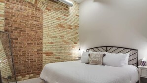 A bedroom with a brick wall, a bed with white bedding, and a nightstand with a lamp.