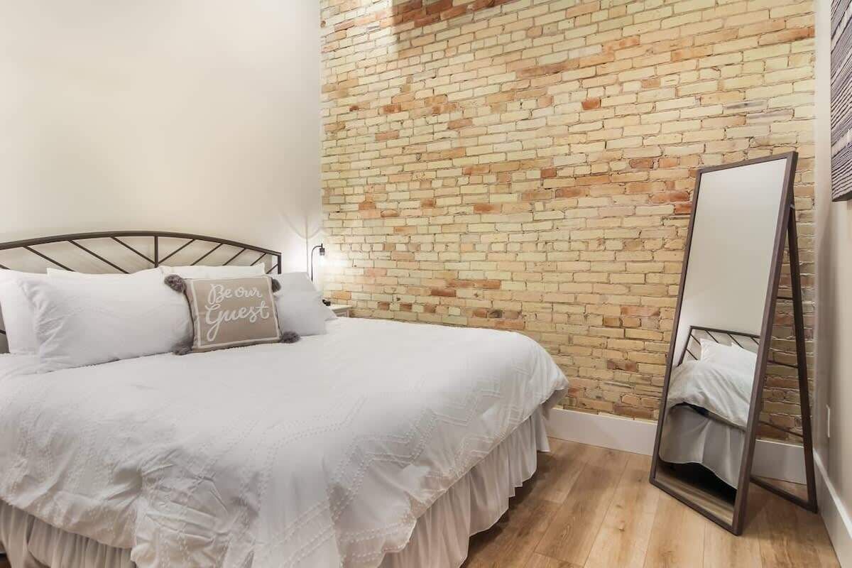 Une chambre avec un mur en briques, un lit avec une literie blanche, un miroir sur la table de chevet et un coussin orné de la phrase « Soyez nos invités ».