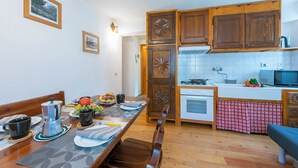 A dining area with a wooden table set for two, a coffee maker, and a fruit bowl. Adjacent is a kitchen with wooden cabinets, a microwave, and a stove. - Miramonti 21 Ski In - Ski Out (Sauze d'Oulx)