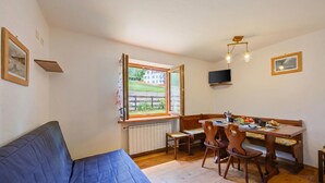 A room with a blue sofa, wooden dining table, chairs, a bench, a window with a view, a radiator, and a TV mounted on the wall. - Miramonti 21 Ski In - Ski Out (Sauze d'Oulx)