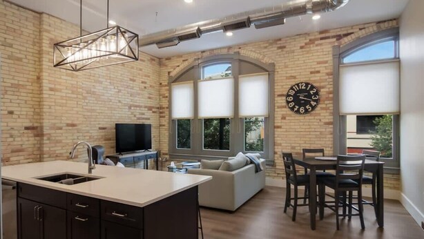 A modern kitchen with a central island, a dining area with a table and chairs, a sofa, and a large window with blinds. - Golden Lofts Unit 3 Downtown Traverse City 2bd/2ba (Traverse City)