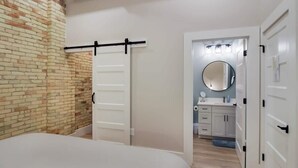A bedroom with a brick wall, a bed, a sliding door, and a bathroom visible through an open door. - Golden Lofts Unit 3 Downtown Traverse City 2bd/2ba (Traverse City)