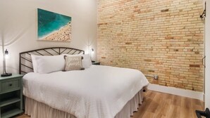 Une chambre avec un mur en briques, un lit recouvert d’une courtepointe blanche, une table de chevet avec une lampe et un tableau représentant une scène de plage.