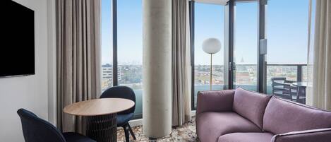 A modern hotel room with a large window, a round table, a sofa, and a TV.