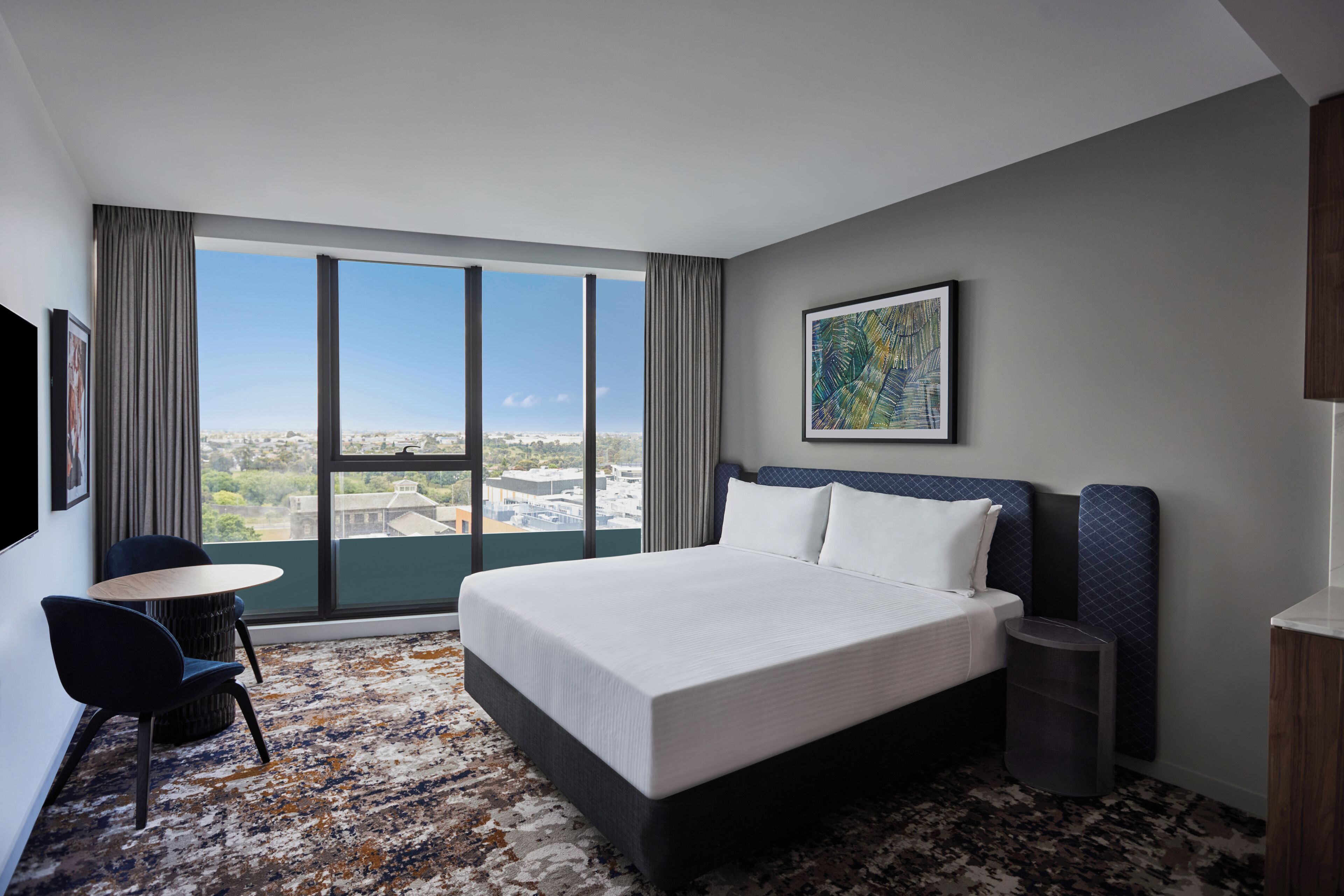 Une chambre d’hôtel avec un grand lit, un bureau, une chaise, une vue sur la ville et une œuvre d’art encadrée au mur.
