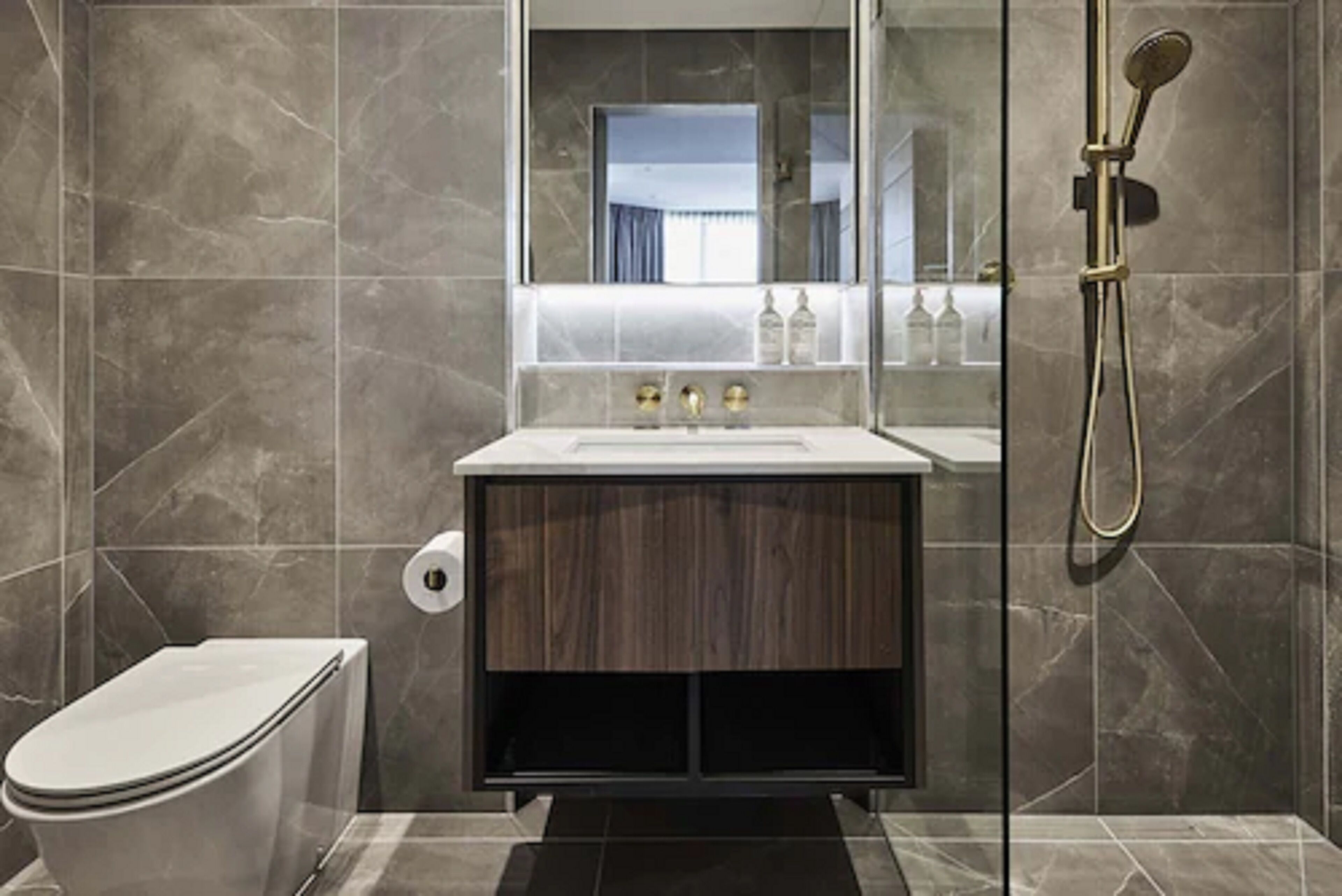 Et moderne badeværelse med en væg belagt med marmorfliser, en trækommode, et hvidt toilet og et bruseområde med glasvægge.