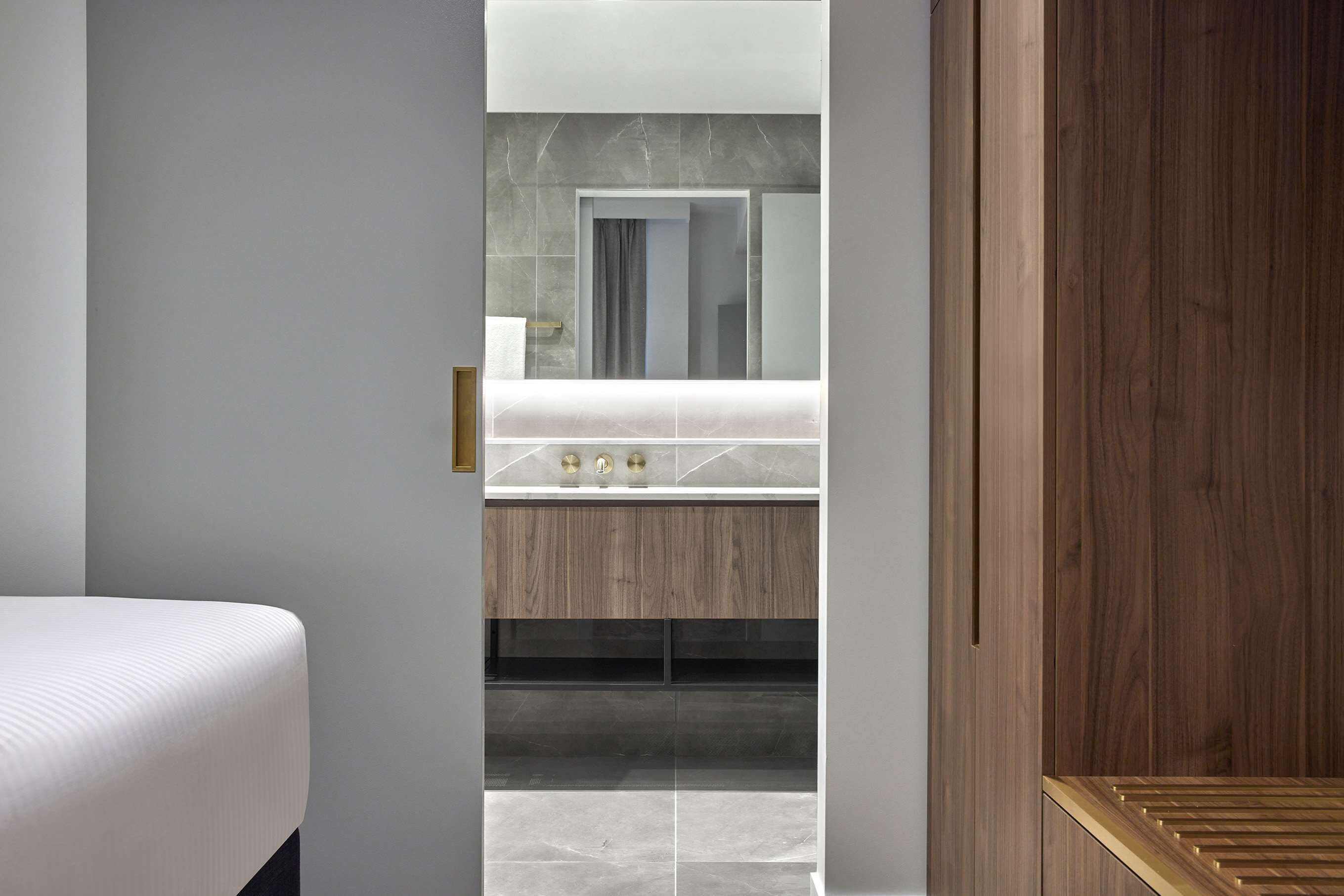 Una habitación de hotel moderna con una cama grande, un armario de madera, un tocador con espejo y un baño visible a través de una puerta abierta.