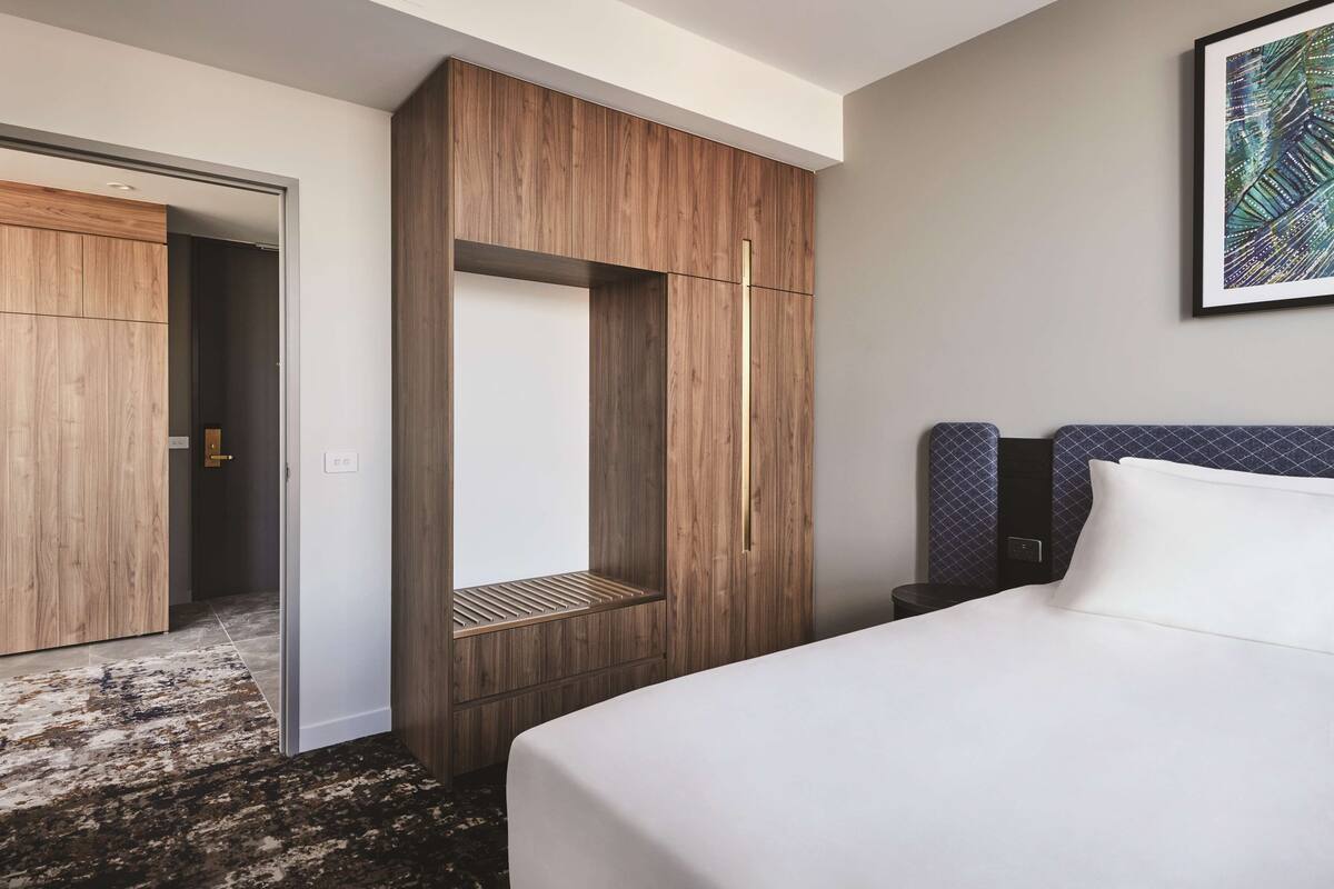 Una habitación de hotel con cama, armario, banco y mesita de noche.