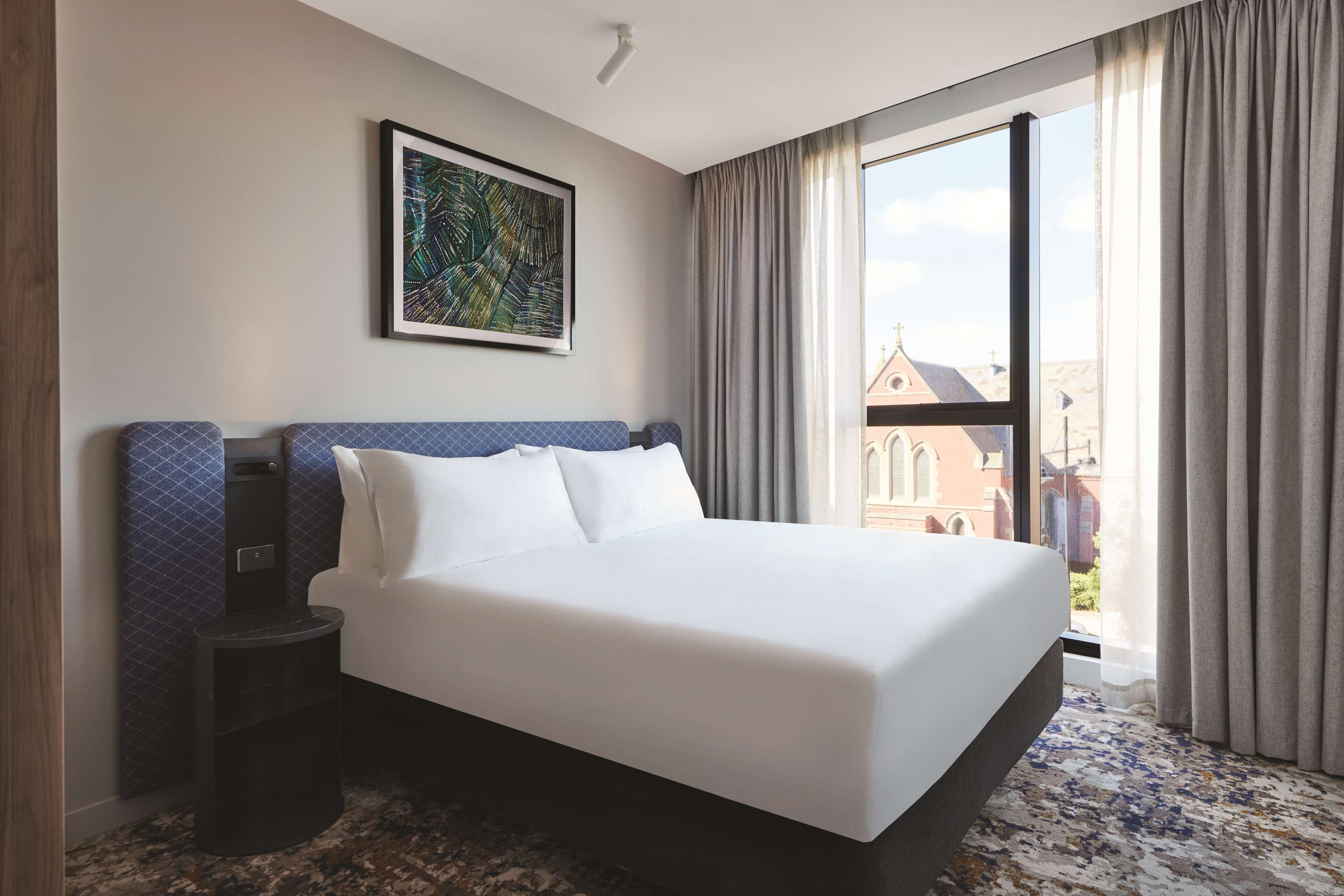 Habitación de hotel con una cama grande, una mesita de noche, un cuadro en la pared y vistas a edificios a través de la ventana.