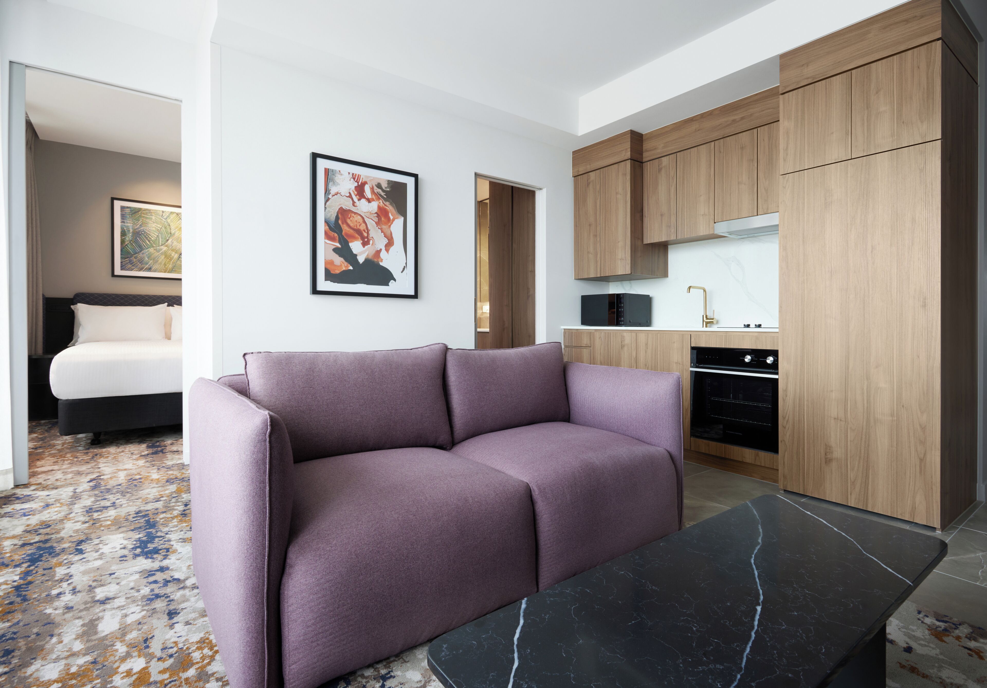 Habitació d'hotel moderna amb un sofà morat, una taula de marbre negre, zona de cuina amb armaris de fusta i un quadre abstracte al mur.