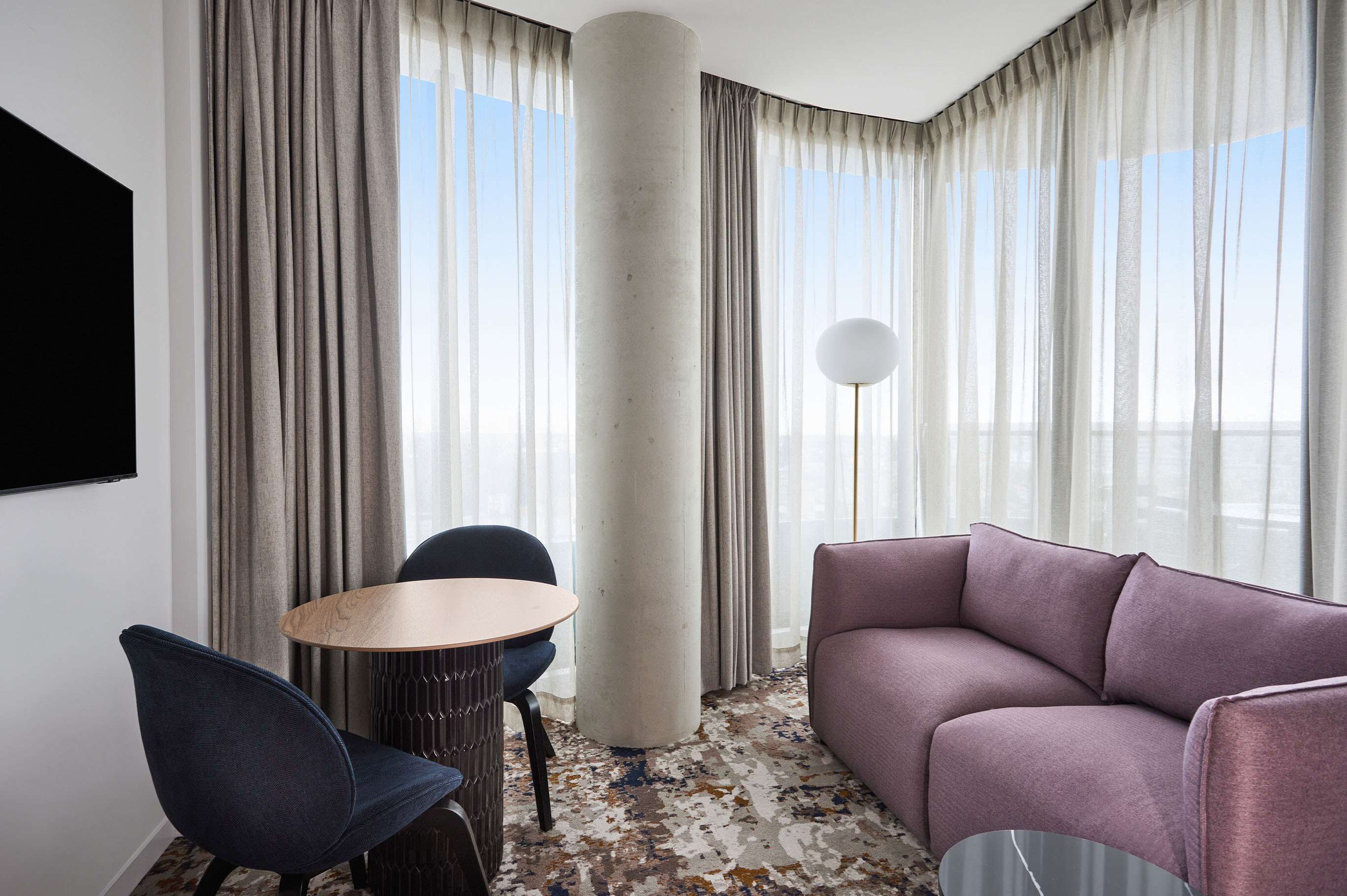 A modern hotel room with a curved ceiling, a large window with sheer curtains, a round table with chairs, a purple sofa, and a floor lamp.