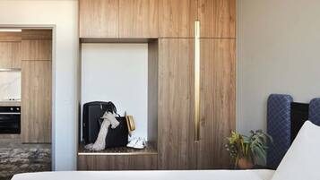 Habitación de hotel con armario empotrado, cama con cabecera blanca, mesita de noche con maleta y sombrero, y planta en maceta.