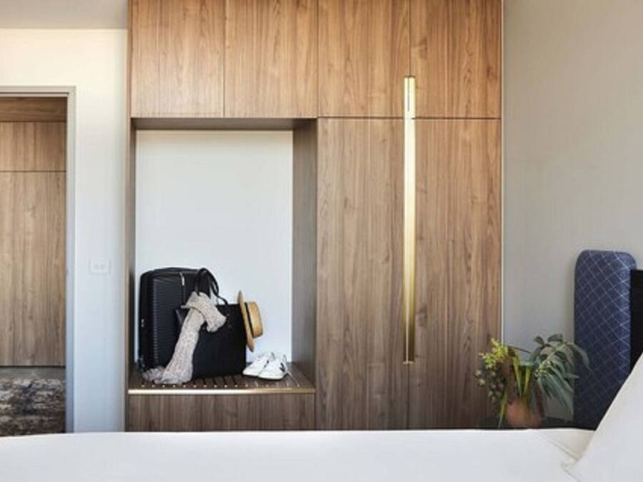 Habitación de hotel con armario empotrado, cama con cabecera blanca, mesita de noche con maleta y sombrero, y planta en maceta.
