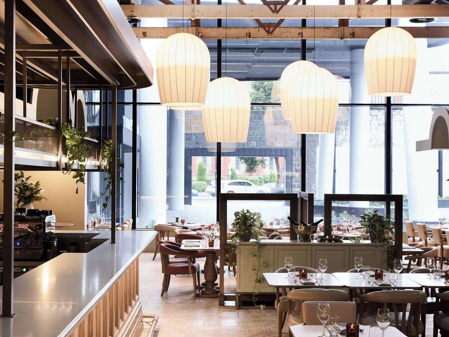 A restaurant with a long counter, multiple tables, hanging light fixtures, and large windows.