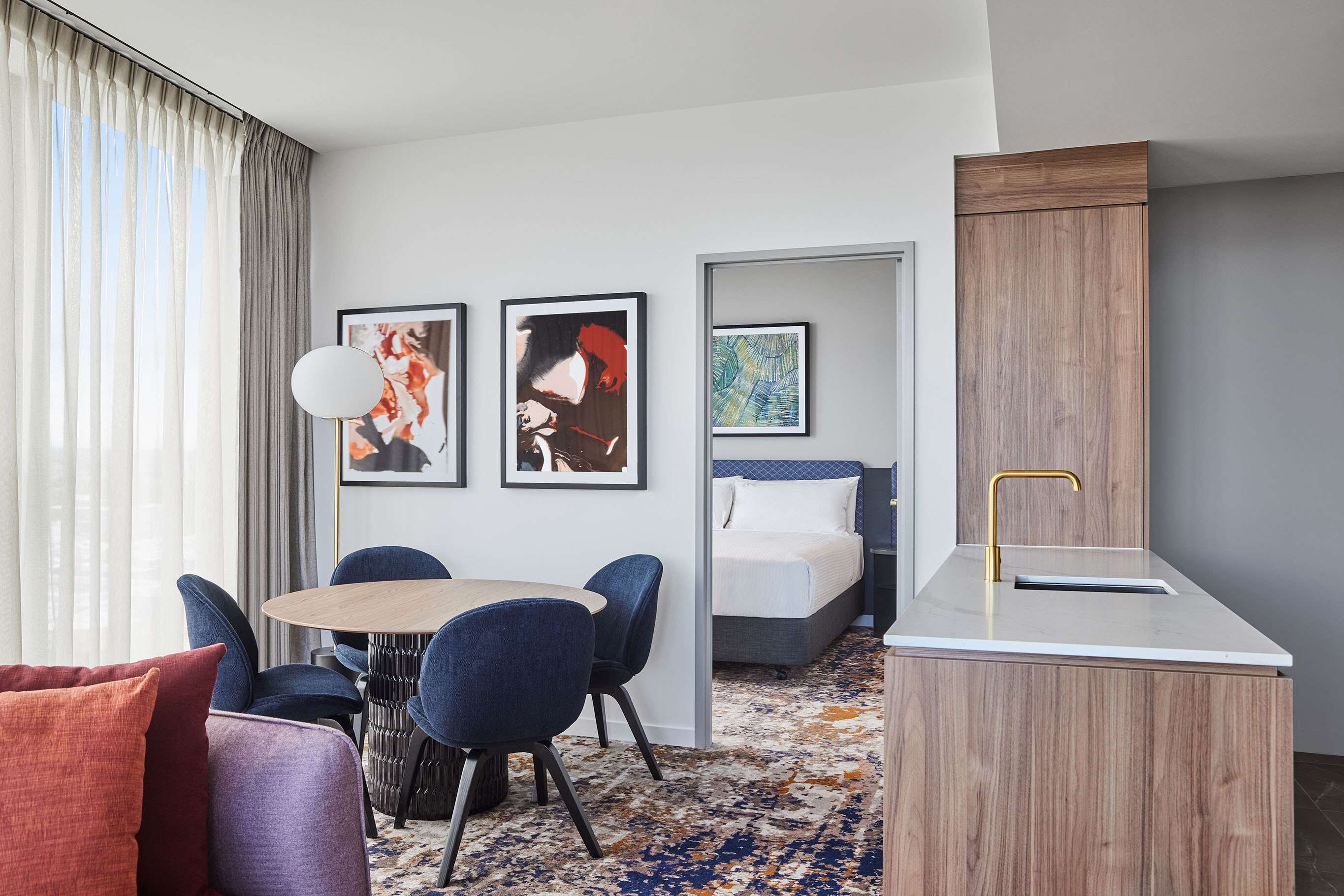 Quarto de hotel moderno com área de refeições, cama com cabeceira e obras de arte nas paredes.
