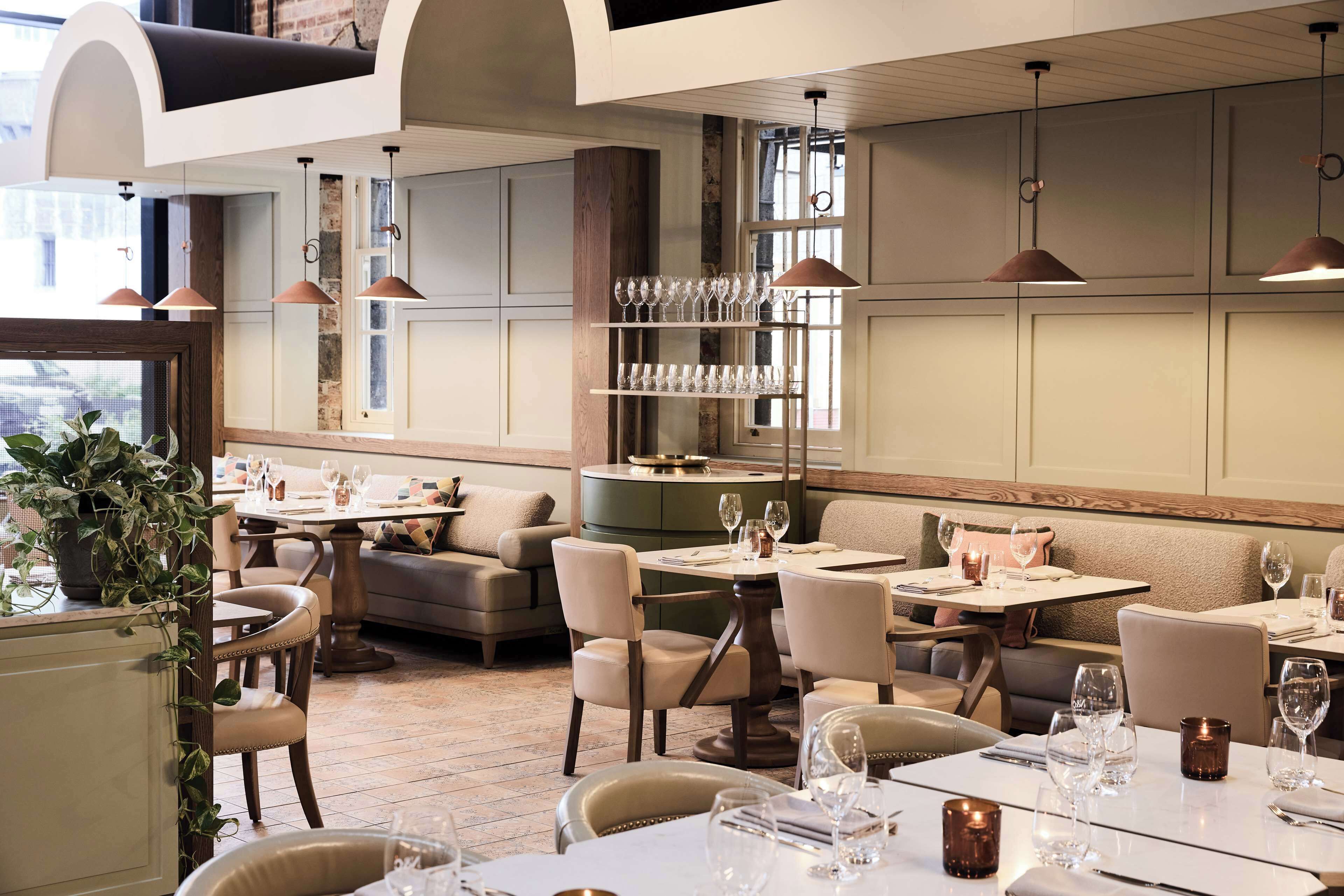A restaurant interior with multiple dining tables, cushioned seating, pendant lighting, and a wine rack.