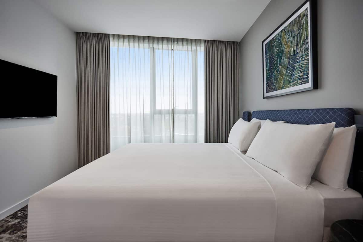 Habitación de hotel con una cama grande, un televisor de pantalla plana en la pared y un ventanal con cortinas translúcidas.
