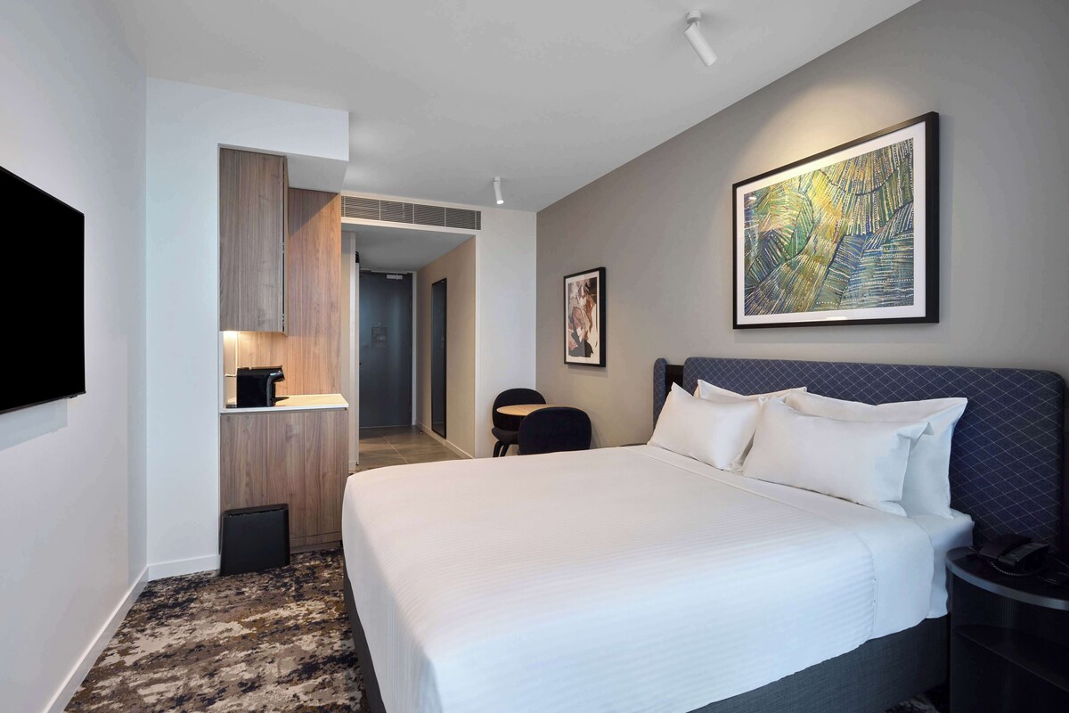 A hotel room with a large bed, a flat-screen TV mounted on the wall, a coffee machine, and a framed artwork above the bed.