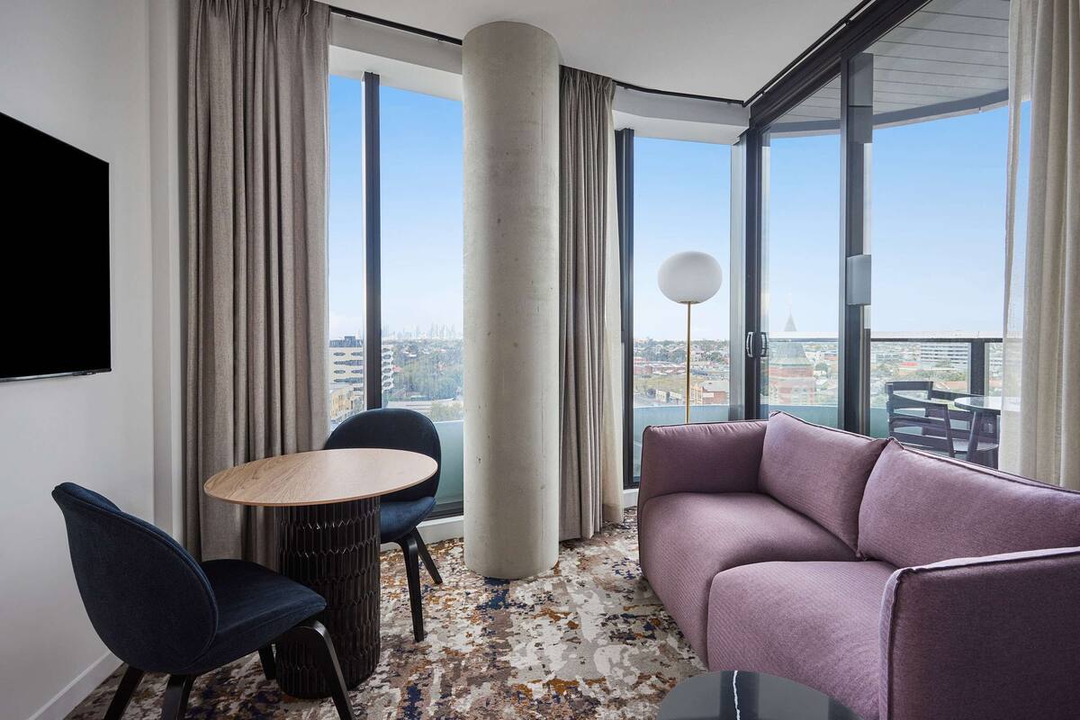 Una habitación de hotel moderna con un ventanal grande, una mesa redonda, un sofá morado y un televisor fijado a la pared.
