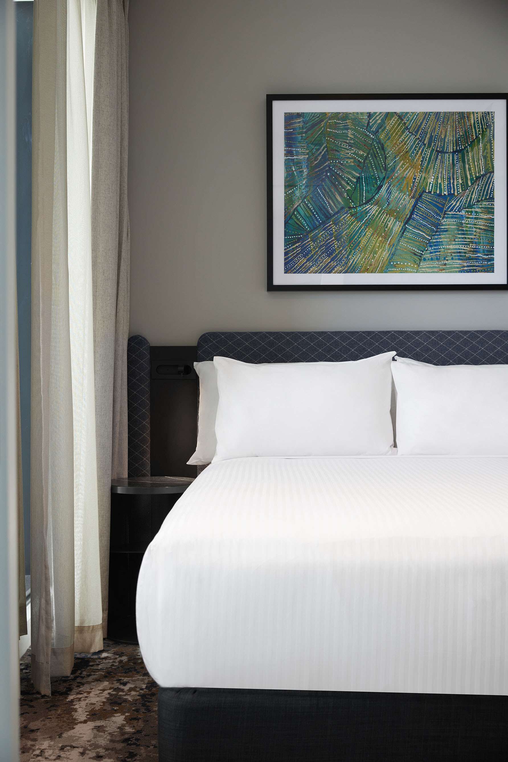 A hotel room with a neatly made bed, a framed abstract artwork on the wall, and a nightstand with a lamp.