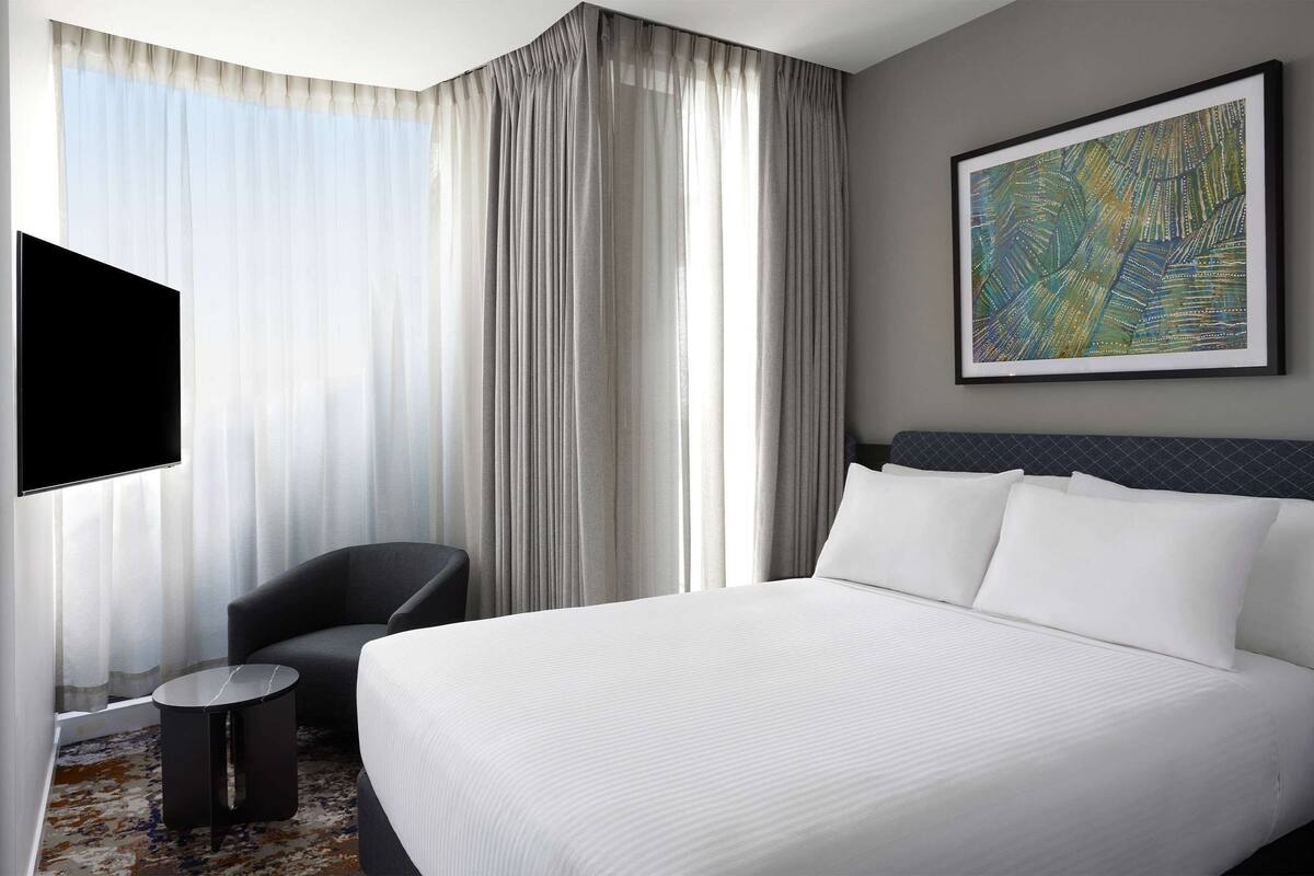 Habitación de hotel con una cama grande, un televisor de pantalla plana montado en la pared, una silla, una mesita redonda y un ventanal con cortinas translúcidas.