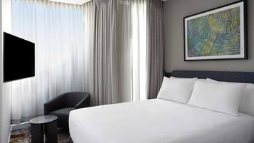 A hotel room with a large bed, a flat-screen TV mounted on the wall, a chair, a small round table, and a large window with sheer curtains.