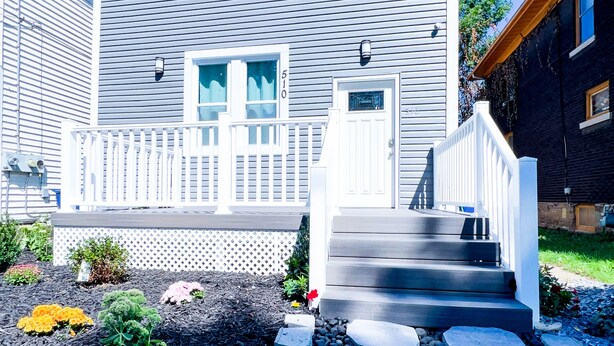 A two-story house with a white front porch, a small deck, and a stone pathway leading to the front door. - The Griffon House (Niagara Falls)