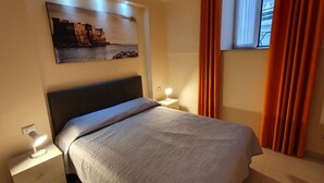 Un dormitorio ordenado con cama, mesitas de noche, una obra de arte en la pared y una ventana con cortinas.