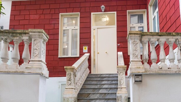 Un edificio rojo con una puerta blanca y dos ventanas, una escalera de piedra con barandilla y un cartelito en la puerta.