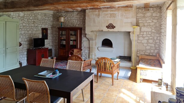 A rustic room with a stone wall, wooden beams, a fireplace, a dining table with chairs, and a bookshelf. - Gîte "Chez Nicole" in the heart of the Loire castles (Restigné)