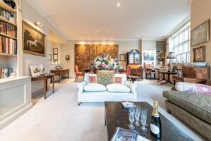 A spacious living room with a sofa, armchairs, a coffee table with glasses and a bottle, and a bookshelf.