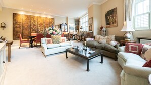Un salon spacieux avec un canapé blanc, une table basse en bois foncé et un fauteuil beige. On y trouve également un coin repas avec une table et des chaises en bois, ainsi qu’un grand tableau accroché au mur.