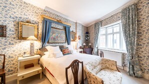 Une chambre à coucher avec un papier peint à motifs floraux, un grand lit, une chaise, une table de chevet, une commode et une fenêtre avec des rideaux.