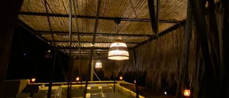Un comedor interior con techo de paja, mesas blancas e iluminación colgante.