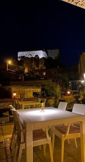 An outdoor dining area with tables and chairs, a stone wall, and a building with arches in the background. - Halici Hani Otel (Ayvacik)