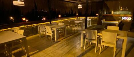 Un patio al aire libre con mesas y sillas, techo de paja y luces colgantes.