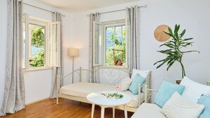 A room with a bed, a small table, two windows with curtains, a plant, and a wall-mounted umbrella.