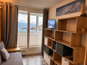 A room with a bed, a TV, wooden shelves, and a balcony with a mountain view.